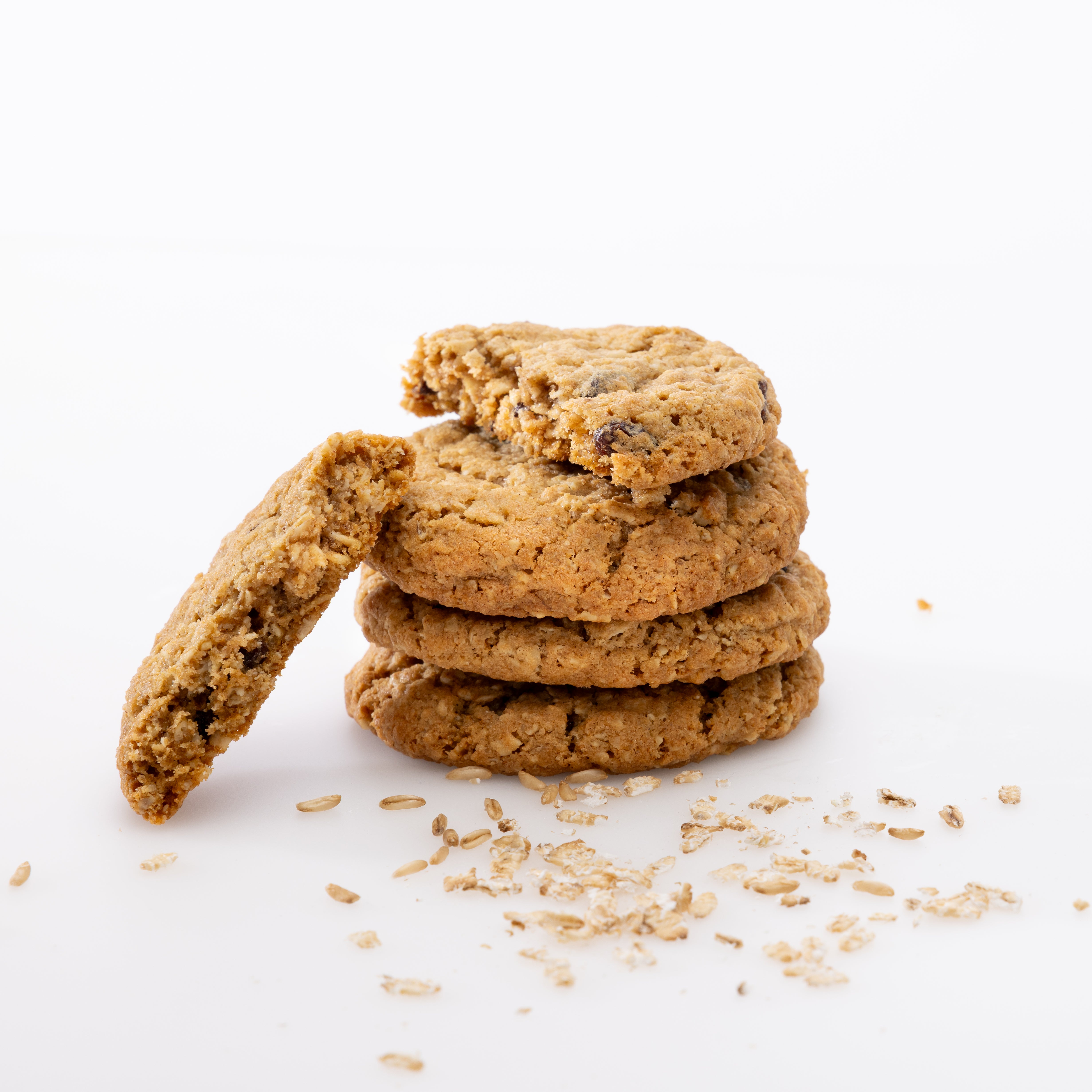 Four golden-brown oatmeal cookies with visible chocolate chips and rolled oats scattered around them on a clean white background.